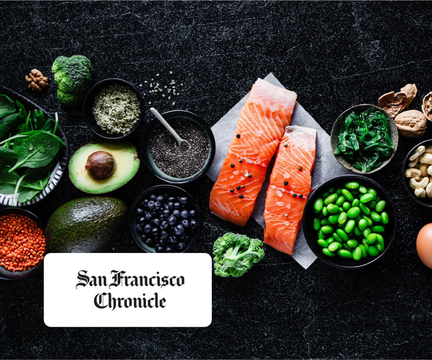 Flat lay of healthy foods on a dark surface, including raw salmon fillets, avocado, spinach, blueberries, edamame, chia seeds, nuts, and lentils, with a “San Francisco Chronicle” label in the foreground.
